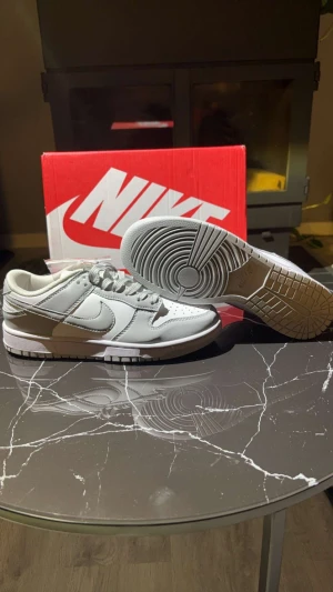 Nike Dunk Low vit/grå sneakers - Nike Dunk Low sneakers i en clean vit och ljusgrå färg. Klassisk siluett med rund tå, snörning och platt sula. Ovandel i skinn och syntet, med perforeringar på tån för extra ventilation. Snyggt broderad Nike-logga på hälen och ikonisk swoosh på sidan.