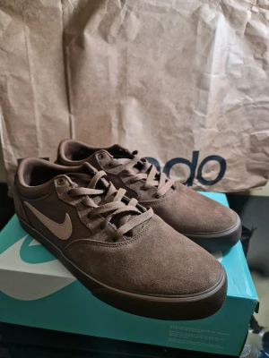 Nike SB Chron 2 bruna sneakers - Nike SB Chron 2 sneakers i brun mocka och textil med klassisk swoosh på sidan. Har endast blivit testad, men va för små. Skorna har snörning, platt sula med mönstrad undersida och rund tå. Insidan har en blå detalj och Nike SB-logga. Perfekt för dig som gillar skate och streetstyle.