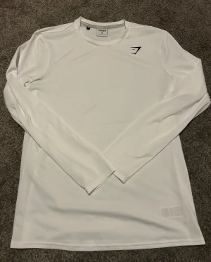 Vit långärmad träningströja Gymshark - Snygg vit långärmad träningströja från Gymshark i storlek M. Tillverkad i 100% återvunnen polyester med ventilerande meshstruktur och rund hals. Perfekt för gymmet eller sport, med diskret logga på bröstet. Enkel och stilren design som passar dig som gillar att hålla det cleant.