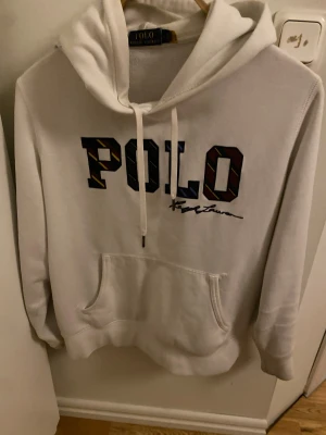 Vit hoodie från Polo Ralph Lauren - Vit hoodie från Polo Ralph Lauren i storlek M. Tröjan har stor POLO-logga med färgglada detaljer på bröstet, snörning vid huvan och en klassisk magficka. Mjuk och skön, perfekt för chill dagar. Materialet är bomull och polyester.