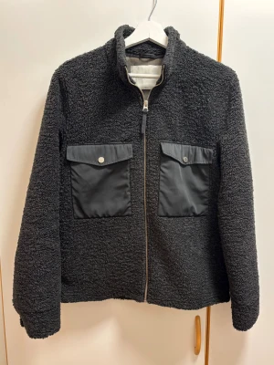 Svart teddyjacka från These Glory Days - Teddyjacka från These Glory Days med två stora fickor framtill och dragkedja. Jackan har hög krage och är tillverkad i ett fluffigt teddy-material som håller dig varm. Perfekt för dig som gillar en stilren och avslappnad look.