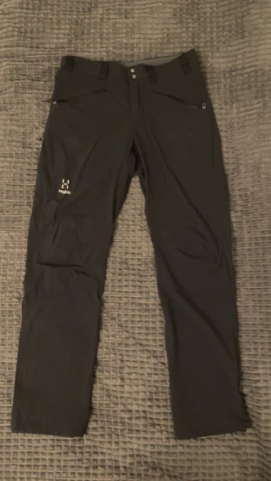 Svarta byxor från Haglöfs, S - Svarta friluftsbyxor från Haglöfs i herrmodell, storlek S. Byxorna har raka ben, flera praktiska fickor med dragkedja och bälteshällor. Materialet är slitstarkt syntet, perfekt för utomhusaktiviteter. Snygg minimalistisk design med diskret Haglöfs-logga på benet.