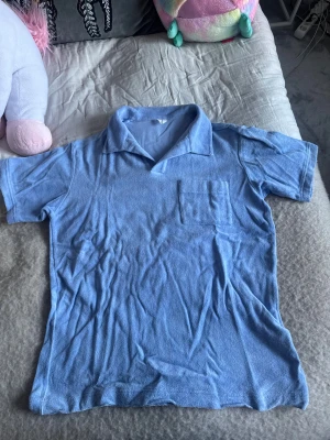 Blå frottéskjorta med krage - Blå skjorta i frottématerial med klassisk krage och korta ärmar. Har en bröstficka på vänster sida och avslappnad passform. 