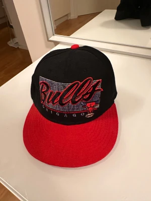  keps svart och röd - Säljer en snygg snapback-keps med broderad logga och text framtill. Kepsen är svart med röd skärm och röd knapp på toppen. Perfekt för dig som gillar basket och streetwear.