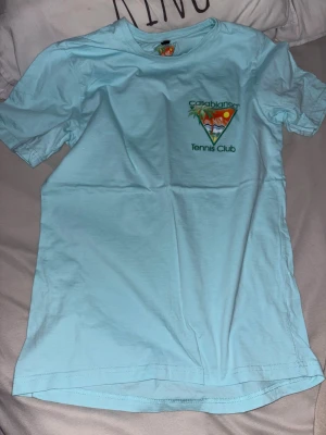 Ljusblå Casablanca Tennis Club t-shirt - Snygg ljusblå t-shirt från Casablanca med stort färgglatt tryck på ryggen och mindre tryck på bröstet. Klassisk passform med rund hals och korta ärmar. Perfekt för dig som gillar sportig och trendig stil.