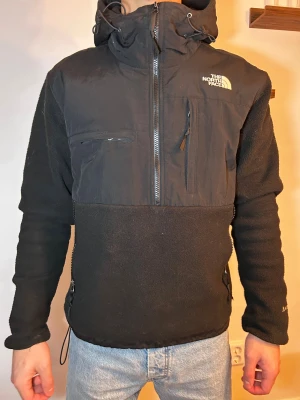 Svart fleecejacka från The North Face - Svart fleecejacka från The North Face med huva och dragkedja framtill. Jackan har flera praktiska fickor med dragkedja och logga på bröstet och ryggen. Materialmix med fleece och syntet, perfekt för kyliga dagar. Snygg och funktionell design med normal passform.