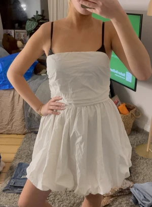 Vit bubblig kortklänning - Supersöt vit kortklänning med bubblig kjol och axelbandslös design. Klänningen har en enkel, stilren look med rynkade detaljer upptill och volym i kjolen. Perfekt för dig som vill sticka ut med en trendig och lekfull stil.