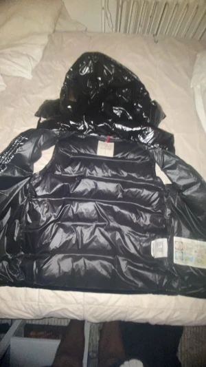 Svart dunväst från Moncler - Snygg svart dunväst från Moncler med glansig finish och huva. Västen har en klassisk quiltad design, dragkedja framtill och detaljer med Moncler-logga. Perfekt för lager-på-lager och ger en sportig vibe till din outfit.