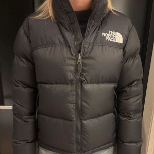 Svart pufferjacka från The North Face - Säljer en svart pufferjacka från The North Face med hög krage och synlig dragkedja framtill. Jackan har ett stilrent, quiltat mönster och logotypen broderad på bröstet och ryggen. Perfekt för kalla dagar och har en normal passform.