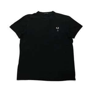 Blå Fred Perry t-shirt med ficka - Blå t-shirt från Fred Perry. På bröstet finns en liten ficka med knapp och det ikoniska Fred Perry-logotypen broderad ovanför. T-shirten är gjord i mjuk bomull och har en clean, stilren look.