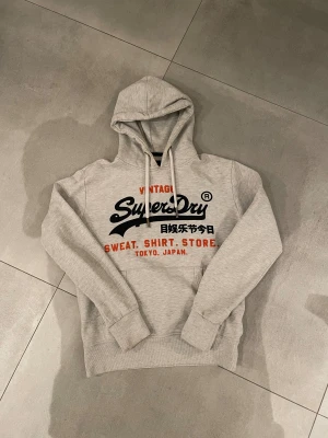 Superdry hoodie grå - Tja Säljer en riktigt snygg ljusgrå hoodie från Superdry med stort svart och orange tryck på bröstet och japanska tecken. Tröjan har huva med snörning, känguruficka och ribbade muddar. Perfekt för dig som gillar den ”nya” grisch stilen och snygga detaljer🤗kan vara den snyggaste hoodien jag sett än så länge🤷‍♂️storlek S och den är i bra skick det enda är att texten är lite sprucken (se bilder) passar dig som är runt 175-185cm lång!! Har du några övriga frågor eller något hör av dig!!!🤩mvh EM_resell🙌🏼