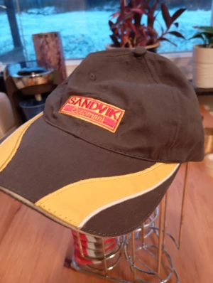 Ny Sandvik keps med gul detalj - Snygg keps från Sandvik Coromant i mörkgrått med gula och vita detaljer på skärmen. Framsidan har ett broderat Sandvik-märke i rött och gult. Perfekt för dig som gillar industriell stil och vill sticka ut med en unik accessoar.