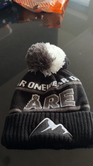 Svart och vit Åre mössa med tofs - Snygg svart och vit mössa från Åre OneWear med stor fluffig tofs på toppen. Stickad design med broderat bergmotiv på uppviket och texten 'ÅRE' framtill. Perfekt för kalla dagar och vinterhäng. Aldrig använd! Pris kan diskuteras! Box följer med 