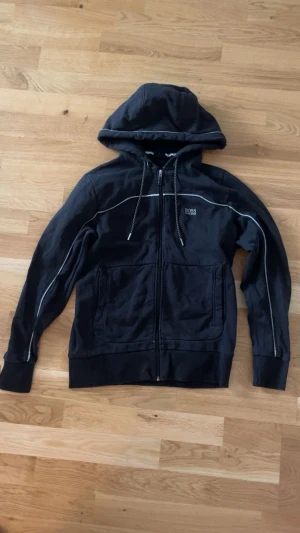 Svart hoodie från Hugo Boss - Svart hoodie från Hugo Boss med vita detaljer längs sömmarna och logga på bröstet. Tröjan har dragkedja, snörning i huvan och två stora fickor framtill. Materialet är mjuk bomull och passformen är normal. Perfekt för chill dagar. Köpte den för 2 år sedan för 2300kr