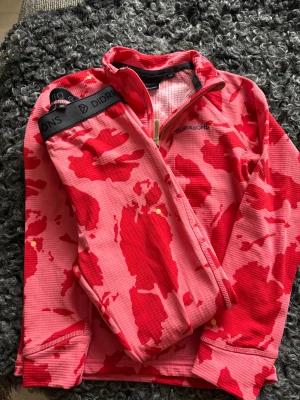 Rosa och röd fleece-tröja Didriksons - Cool långärmad lite tjockare underställströja med tillhörande byxor från Didriksons i ”rosa” och rött med camouflagemönster. Tröjan har half zip med gul detalj på dragkedjan och broderad logga på bröstet. Knappt använda. Perfekt för vinterväder eller chill dagar när du vill vara både bekväm och snygg.