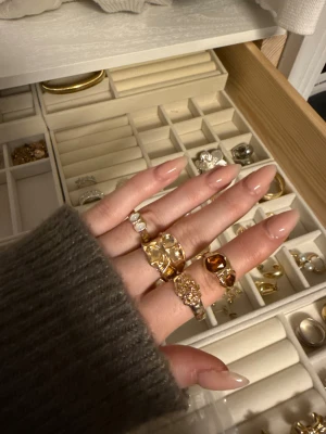 Fyra chunky guldringar med stenar - Säljer ett set med fyra chunky guldfärgade ringar i olika former. En ring har två vita pärlor, en har två bruna stenar, en har en abstrakt yta och en har ett detaljerat mönster. Perfekt för dig som gillar statement-smycken och att mixa olika stilar.