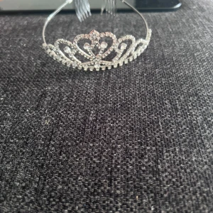 Glittrigt diadem med strass - Snyggt diadem i silverfärgad metall med glittrande strass-stenar i ett elegant, kronliknande mönster. Perfekt för att ge din look extra bling och prinsesskänsla. Diademet har kammar på sidorna för att sitta stadigt i håret.