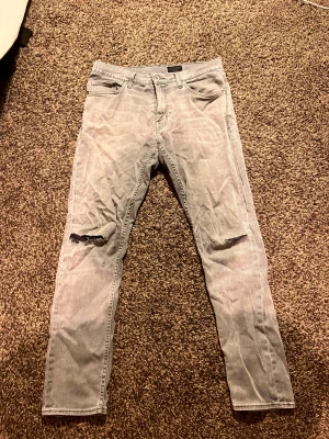 Grå Tiger of Sweden jeans Pistolero - Säljer ett par grå jeans från Tiger of Sweden, modell Pistolero. De har slim passform, klassisk femficksdesign och slitna detaljer vid knäna för en cool look. Materialet är jeans och färgen är ljusgrå. Perfekt för dig som gillar stilrena och trendiga byxor.