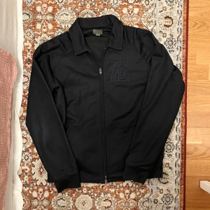 Svart overshirt från Acne Studios - Svart overshirt från Acne Studios med broderad logga på bröstet. Jackan har dragkedja framtill, klassisk krage och långa ärmar. Materialet är syntet med meshfoder på insidan för extra komfort. Perfekt för dig som gillar stilrena och cleana plagg. Plagget är i fint skick förut att lappen är påväg att åka av, men så har den suttit i cirka 4 år nu så va inte oroliga att den ska åka av