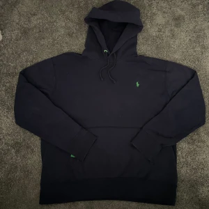 Mörkblå hoodie från Polo Ralph Lauren - Mörkblå hoodie från Polo Ralph Lauren med klassisk grön logga broderad på bröstet. Tröjan har huva med snörning och långa ärmar. Skriv vid frågor