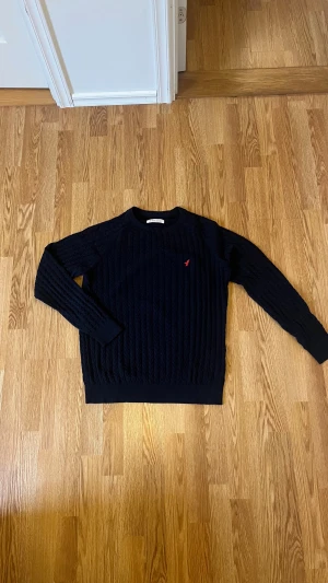Mörkblå stickad tröja från Pier One - Snygg mörkblå stickad tröja från Pier One med kabelstickat mönster och en liten röd broderad fågel på bröstet. Rund hals och ribbade muddar vid ärmslut och nederkant. Perfekt för dig som gillar klassisk stil med en twist.