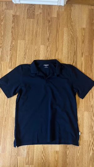Marinblå pikétröja Jack & Jones M - Snygg marinblå pikétröja från Jack & Jones Essentials i storlek M. Klassisk krage och korta ärmar, med diskret broderad logga på bröstet. Tröjan är gjord i mjuk bomull som känns skön mot huden och passar perfekt till jeans eller shorts.