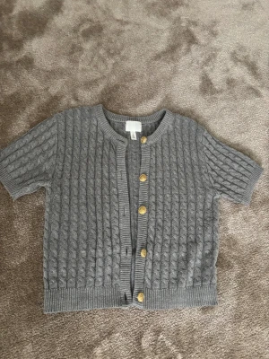 grå kofta med guld knappar - Superfin grå kofta i tjockare kabelstickad kvalitet. Korta ärmar och rund halsringning. Guldfärgade knappar framtill som ger ett lite mer unikt och stilrent uttryck. Modellen är något cropped och sitter snyggt både öppen och stängd. Säljer då den inte kommer til användning 
