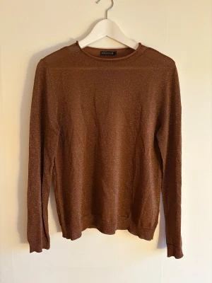 Brun glittrig tröja från Stockh LM - Snygg brun långärmad tröja från Stockh LM, modell Carrie Sweater. Tröjan har ett glittrigt material som ger en cool shimmer-effekt och är i en normal passform. Perfekt för dig som gillar att sticka ut med lite extra shine.