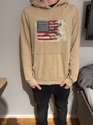 Ralph Lauren hoodie med  amerikansk flagga - Säljer denna feta hoodie med tryck av en amerikansk flagga med vintage-look och textdetaljer. Hoodien är tillverkad i mjukt bomullsmaterial och har ribbade muddar vid ärmslut och nederkant.