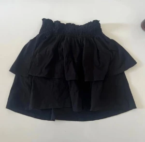 Svart volangkjol från Gina Tricot - Supersöt svart kortkjol från Gina Tricot med bred smock i midjan och dubbla volanger. Kjolen är gjord i mjuk bomull och har en flowy känsla som ger en cool och avslappnad vibe. Perfekt att styla med en crop top eller hoodie.