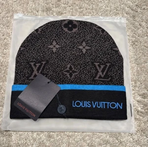 Mössa - Svart och mörkgrå mössa  med klassiskt monogrammönster och blå kant. broderad i blått framtill. Snygg och stilren design som sticker ut, perfekt för dig som gillar exklusiva accessoarer. Materialet är stickat och mjukt.