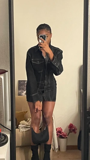 Svart jeansklänning med lång ärm - Säljer en svart jeansklänning med markerade vita sömmar och långa ärmar. Klänningen har skjortkrage, knappar framtill och fickdetaljer på bröstet. Kort modell som sitter snyggt och har en cool street vibe. Perfekt att styla med boots och liten väska.