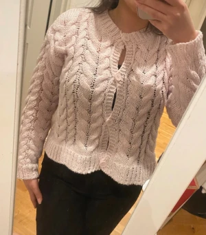 Ljusrosa stickad kofta med knappar - Säljer en ljusrosa stickad kofta med grova flätstickade detaljer och rund hals. Koftan har långa ärmar och stängs med fyra matchande knappar framtill. Perfekt för dig som gillar mysiga och trendiga plagg med lite vintage-vibe.