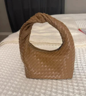 Flätad brun handväska från Gina Tricot - Snygg brun handväska från Gina Tricot med flätad design och knut på handtaget. Väskan har dragkedja upptill och är gjord i syntetmaterial. Perfekt storlek för det viktigaste och ger en trendig touch till din outfit.