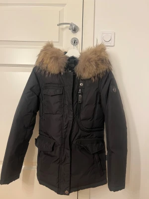 Svart parkajacka med päls från Hollies - Svart parkajacka från Hollies med äkta päls. Jackan har flera fickor med lock och dragkedja framtill, samt en snygg badge på ärmen. Perfekt för kalla dagar med sin långa passform och varma material. Ny pris 4000kr. 