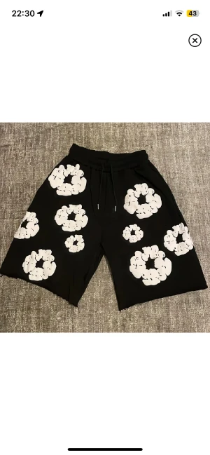 Svarta shorts med vita blommor Denim Tears - Svarta shorts från Denim Tears med stora vita blommor tryckta över hela plagget. Modellen har midwaist passform, elastisk midja med snörning och är gjorda i mjuk bomull. Perfekt för dig som vill sticka ut med ett unikt mönster och streetwear-vibe.