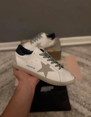 Golden Goose vit sneakers - Golden Goose sneakers i vitt skinn med grå snörning och en stor grå stjärna på sidan. Hälpartiet är svart och sulan är platt och beige. Snygg och stilren modell med GGDB/STAR-tryck på sidan. Levereras med originalkartong och dustbag. Priset går att diskutera