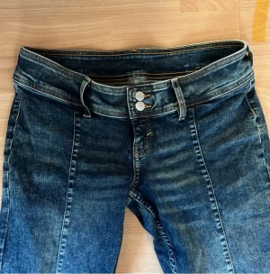 Blå bootcut jeans med låg midja - Snygga blå jeans med bootcut och låg midja, markerade sömmar framtill och dubbla knappar i midjan. Klassisk denim-look med lätt slitning och orange kontrastsömmar. Perfekt för dig som gillar retrovibbar och vill ha ett par jeans som sticker ut.