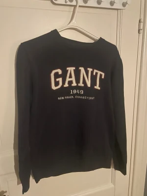 Svart GANT sweatshirt med tryck i storlek M - Svart sweatshirt från GANT med stort broderat tryck på bröstet i vitt och rött. Rund halsringning och långa ärmar. Mjuk och skön bomullskvalitet, perfekt för chill dagar. Klassisk college-look med ribbade muddar vid ärmslut och nederkant.