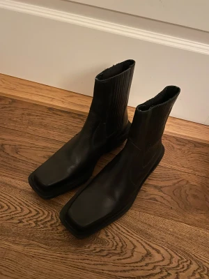 Alohas svarta boots med fyrkantig tå - Säljer ett par svarta boots i skinn med fyrkantig tå och låg klack. Snygg minimalistisk design med ribbade detaljer på skaftet och platt sula. Perfekt för dig som gillar stilrena och moderna skor med edge.