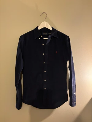 Mörkblå manchesterskjorta Polo Ralph Lauren - Slim fit skjorta i mörkblå manchester från Polo Ralph Lauren. Klassisk krage, vita knappar och röd broderad logga på bröstet. Långärmad och perfekt för dig som gillar stilren och tidlös design.