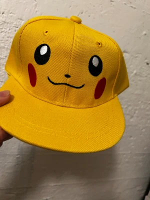 Gul Pikachu keps med tryck - Snygg gul keps med Pikachu-ansikte tryckt framtill. Kepsen har en klassisk form med böjd skärm och justerbar snapback baktill. Materialet är slitstark bomull och färgen är riktigt klar gul med svarta och röda detaljer. 3-7 år 