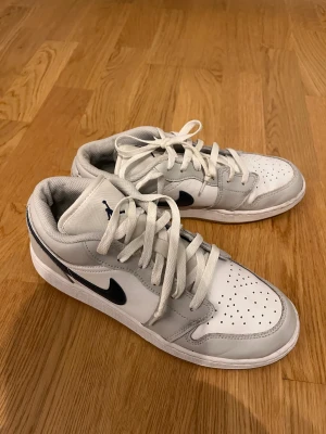 Nike Jordan 1 Low vit/marinblå sneakers - Nike Jordan 1 Low sneakers i vit och grått med marinblåa detaljer. Klassisk design med perforerad tå, snörning och rund tå. Skorna har en svart swoosh på sidan och Jordan-logga på hälen. Tillverkade i skinn och syntetmaterial för en clean och sportig look.