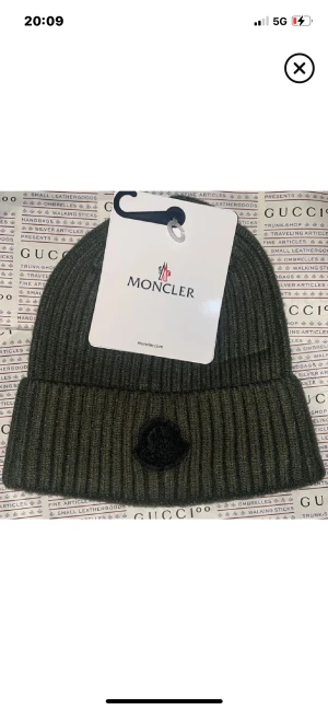Ribbad grön mössa från Moncler - Snygg ribbad mössa från Moncler i mörkgrön färg med uppvikt kant och svart broderad logga framtill. Perfekt för dig som vill ha en clean och stilren look med exklusiv känsla. Materialet är mjukt och värmande, passar perfekt när det är kallt ute.