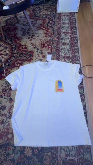 Vit t-shirt från Casablanca - Säljer en clean vit t-shirt från Casablanca med färggrafiskt tryck fram och bak. Trycket har en cool fönsterdesign i gult, blått och rött med texten 'Casa Blanca'. T-shirten har rund hals och korta ärmar, perfekt för dig som gillar stilrena vibes och grafiska prints.