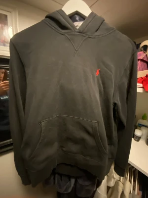 Svart hoodie från Polo Ralph Lauren - Svart hoodie från Polo Ralph Lauren med klassisk röd logobrodyr på bröstet. Tröjan har huva, känguruficka och långa ärmar. Mjuk bomullskvalitet som är skön att bära. Perfekt för dig som gillar stilren och enkel design.