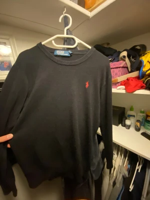 Svart stickad tröja Polo Ralph Lauren - Svart stickad tröja från Polo Ralph Lauren med klassisk röd broderad logga på bröstet. Rund halsringning och långa ärmar. Perfekt för dig som gillar stilrena och tidlösa plagg med en touch av lyx.
