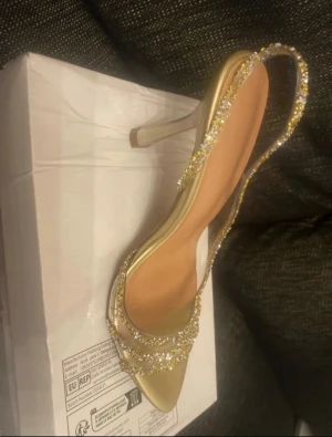 Glittriga guld klackar  - Säljer ett par eleganta pumps i guld med spetsig tå och hög klack. Skon har glittriga detaljer med strass längs remmarna och runt hälen, vilket ger en lyxig och festlig vibe. Perfekt för dig som vill sticka ut med stil och glans.