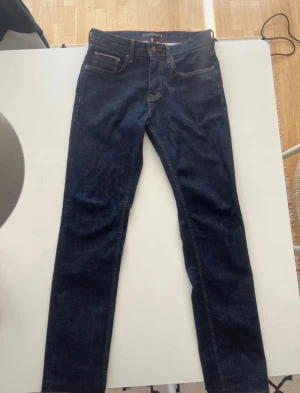 Tommy Hilfiger Denton Stretch Jeans - Snygga mörkblå jeans från Tommy Hilfiger, modell Denton Stretch Straight Fit. Klassisk femficksdesign med bruna sömmar och läderpatch bak. Materialet är 96% bomull och 4% elastan för skön stretch. Diskret Tommy Hilfiger-logga på bakfickan.