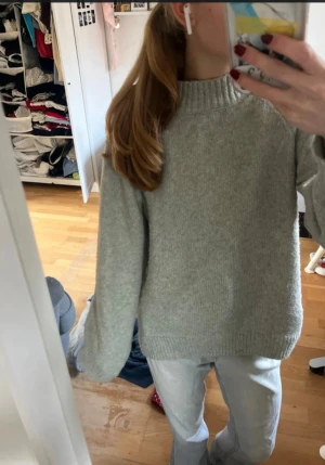 Grå stickad tröja med hög krage - Mysig grå stickad tröja med hög ribbad krage och långa ärmar. Tröjan har en loose passform och är perfekt för kyliga dagar. Enkel och stilren design som funkar till jeans eller andra favoritplagg.